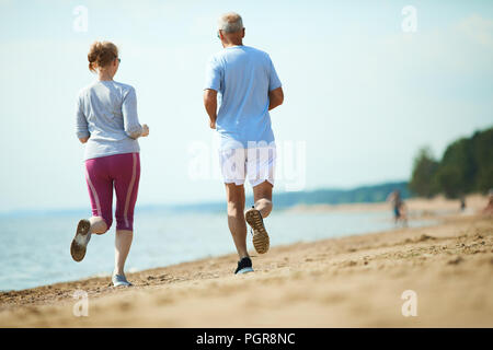 Back view of senior couple in activewear running along coastline on sandy beach Stock Photo