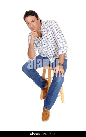 A tall handsome man sitting on a bar chair in a checkered shirt and jeans with one hand on his chin, isolated for white background Stock Photo