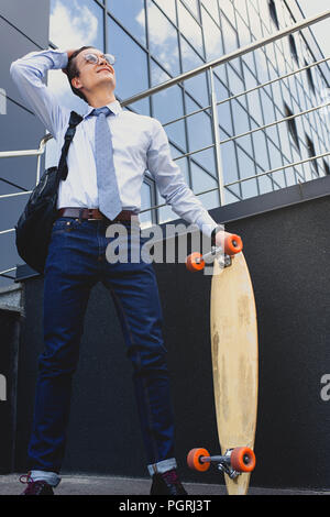 smiling young businessman in eyeglasses standing with longboard and looking away Stock Photo