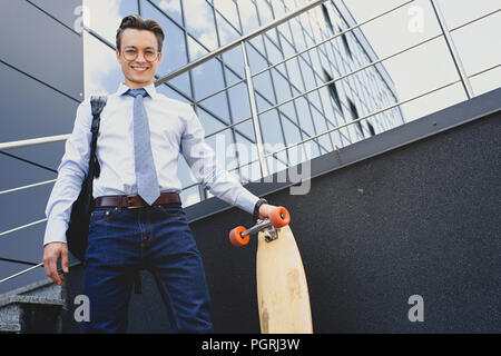 low angle view of handsome young businessman in eyeglasses standing with longboard and smiling at camera Stock Photo
