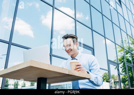 low angle view of smiling young freelancer holding paper cup and using laptop outside modern building Stock Photo