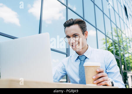 handsome smiling young businessman in eyeglasses using laptop and holding paper cup outside modern building Stock Photo