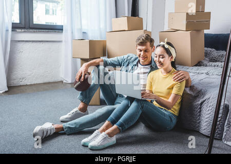 happy young couple sitting on floor of bedroom and using laptop while moving into new home Stock Photo