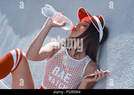 Young woman on roller skates and helmet drinking water. Stylish beautiful girl in shorts and a purple shirt rollerblading. Hipster girl resting after  Stock Photo