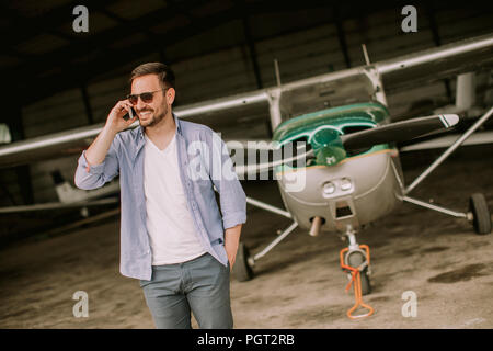 Young handsome young pilot checking airplane in the hangar and using mobile phone Stock Photo