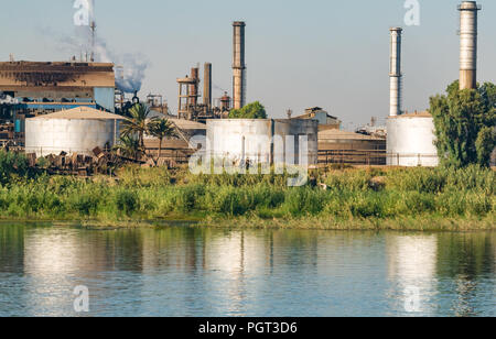 Industrial site on riverbank with steam towers and storage containers reflected in water, Nile River, Egypt, Africa Stock Photo