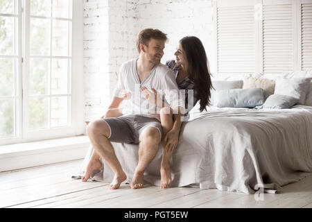 Happy couple sitting on bed hugging looking in the eyes Stock Photo