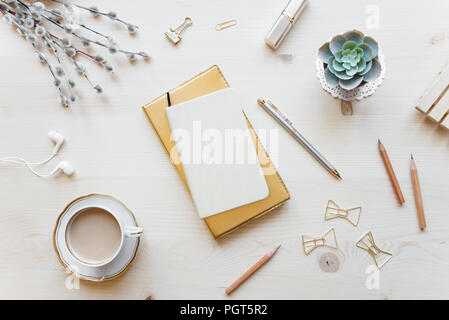 Female home office desk table in golden tones. Workspace with notebooks, cup of coffe and decorations on light wooden background with copy-space. Flat Stock Photo