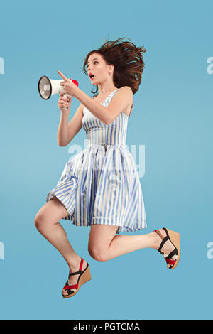 Beautiful young woman jumping with megaphone isolated over blue background. Runnin girl in motion or movement. Human emotions and facial expressions concept Stock Photo