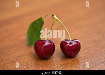 Two red ripe cherries with stems and a leaf on a wood table Stock Photo