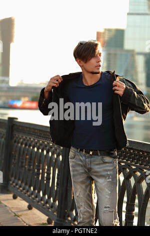 Thinking handsome young man walking in fashion jeans and dressing the black jacket on city center background. Stock Photo