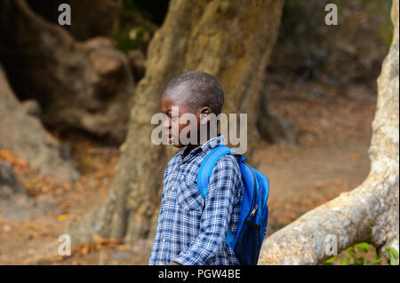 SOGA, GUINEA BISSAU - MAY 5, 2017: Unidentified local little boy in plaid shirt with backpack walks along the street in a village of the Soga island.  Stock Photo