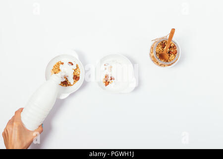 Making healthy breakfast process, top view. Women's hand pouring milk or yogurt into bowl with homemade granola. Stock Photo