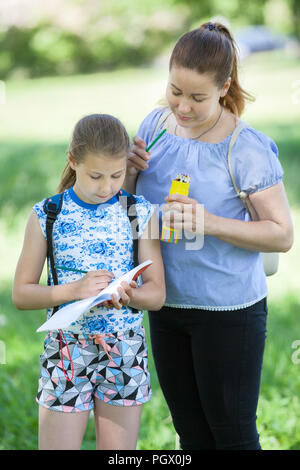 Mother looks in sketchbook, holding color pencils and help daughter to draw at nature Stock Photo