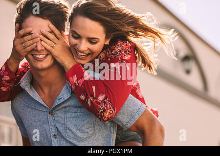 Smiling woman piggy riding on man closing his eyes with her hands. Smiling man carrying woman on his back walking outdoors. Stock Photo