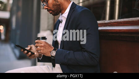 African businessman sitting outside with coffee and using mobile phone. Man in suit relaxing outdoors by the road texting on his cellphone and drinkin Stock Photo