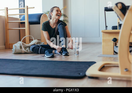 Smiling woman wearing her shoes after workout sitting at the gym. Woman sitting on floor at a pilates gym with her gym bag tying her shoelaces. Stock Photo
