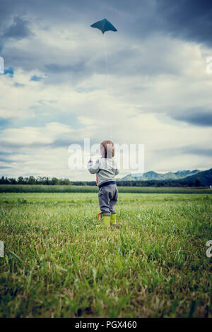 View from behind of a cute young child in gumboots standing flying a kite in a grassy green field standing holding the string watching it soar in the  Stock Photo