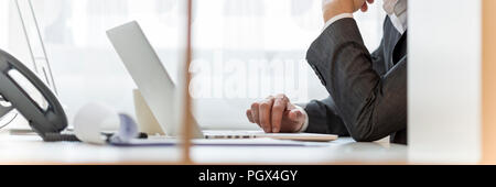 Businessman sitting at his office desk working on laptop computer with paperwork on table, wide cropped view. Stock Photo