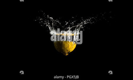 Water splashing from the surface of the liquid in a spray of droplets as a halved lemon is dropped into the water from above on a dark background with Stock Photo