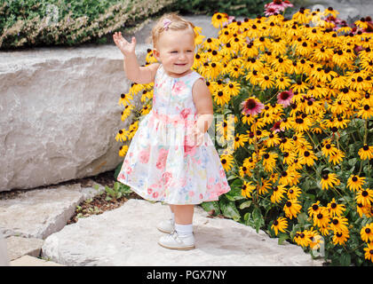 Cute adorable white Caucasian baby girl child in white dress standing among yellow flowers outside in garden park looking away, lifestyle happy childh Stock Photo