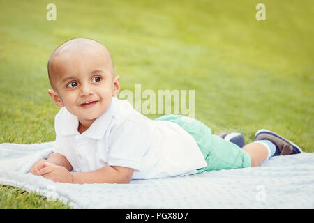 Portrait of cute adorable little indian South Asian or Middle Eastern infant boy in white shirt laying on ground in park outside on bright summer day  Stock Photo