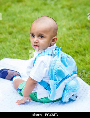 Portrait of cute adorable little indian mixed race infant boy in white shirt sitting on ground with blue backpack schoolbag in park field green grass  Stock Photo