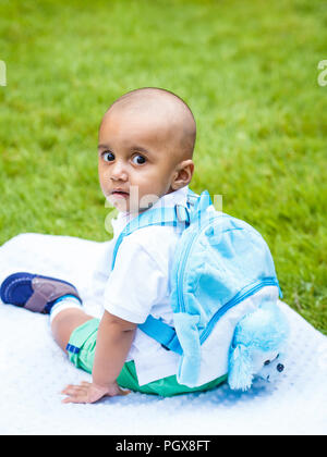 Portrait of cute adorable little indian mixed race infant boy in white shirt sitting on ground with blue backpack schoolbag in park field green grass  Stock Photo