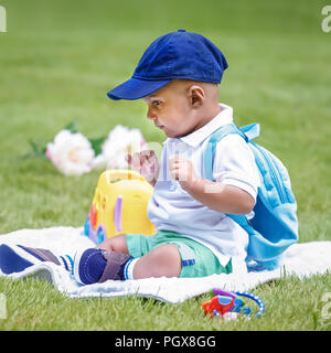 Portrait of cute adorable little indian mixed race infant boy in white shirt sitting on ground with blue backpack schoolbag and toys in park field gre Stock Photo