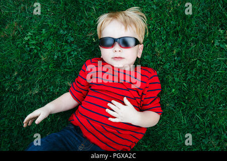 Close up portrait of funny cute adorable white Caucasian toddler child boy with blond hair in red pullover sunglasses lying on green grass. View from  Stock Photo