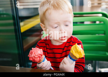 Portrait of cute adorable white Caucasian funny blond child boy holding two ice cream in waffle cone, trying to choose one, looking surprised puzzled, Stock Photo