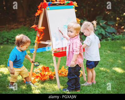 Group of three white Caucasian toddler children kids boys and girl standing outside in summer autumn park by drawing easel with markers, playing study Stock Photo