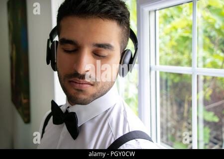 Man in tuxedo with bow tie, stands next to window with headphones and eyes closed Stock Photo