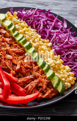 Slow cooker pulled pork rancho salad with arranged rows of thinly sliced red cabbage, corn, sweet pepper, cucumber and chopped jalapeno on a black pla Stock Photo