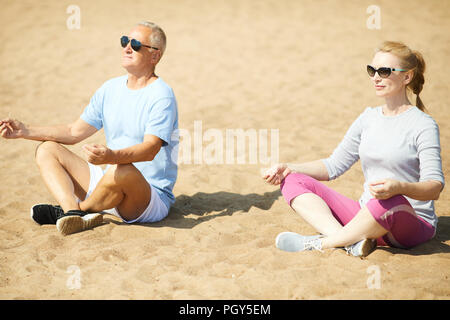 Contemporary senior couple in activewear and sunglasses sitting on sand in pose of lotus after workout Stock Photo
