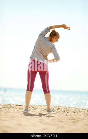 Rear view of active mature woman standing on beach and side-bending by waterside Stock Photo