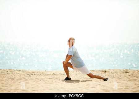 Senior man in activewear stretching his legs while exercising on sandy beach by waterside Stock Photo