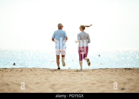 Happy energetic aged couple in activewear running towards waterside while training on beach Stock Photo