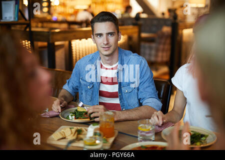 Young man siting by served table among his friends, eating lunch and talking to one of buddies Stock Photo