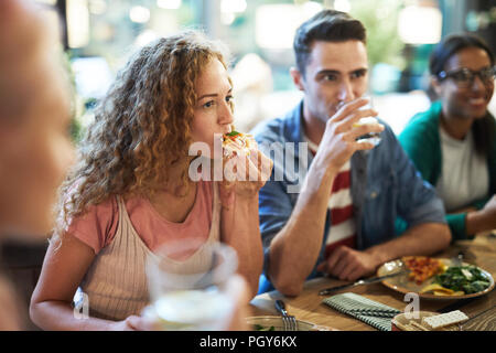 Hungry casual girl eating pizza by dinner among her friends and listening to one of them during chat Stock Photo