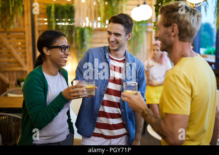 Two laughing guys and girl with glasses of lemonade making toast or just having chat in cafe Stock Photo