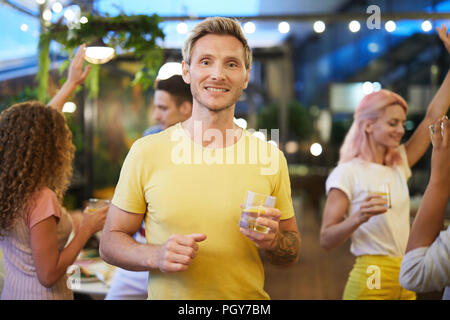 Happy guy in yellow t-shirt holding glass of drink while dancing among his friends at party Stock Photo