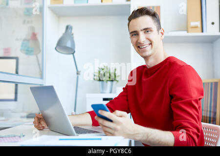 Smiling young man in casualwear using modern gadgets while sitting by desk in office Stock Photo