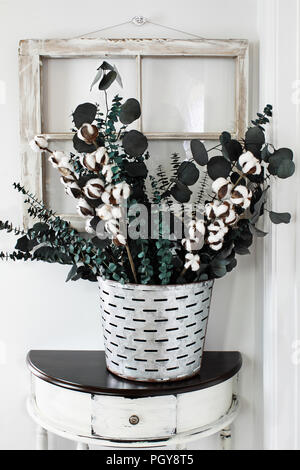 Note to reviewer. Items are all generic. Old olive bucket filled with eucalyptus and cotton stem arrangement on a farmhouse table in front of vintage  Stock Photo