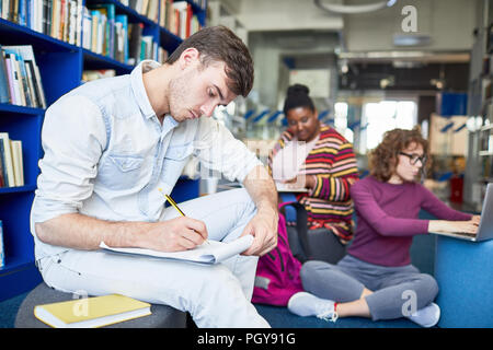 Concentrated male student writing report while sitting in library with other multiethnic students in the background Stock Photo