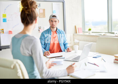 Confident handsome guy in cap discussing university project with groupmate while they sitting at table and finding solution Stock Photo