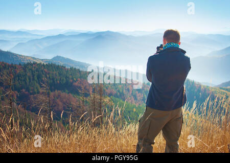 One young man standing with his back turned, holding camera and making photos of a mountain range covered in red, orange, yellow deciduous forest unde Stock Photo