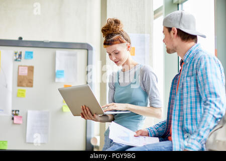 Serious modern students sitting on window sill and using laptop while discussing project in classroom Stock Photo