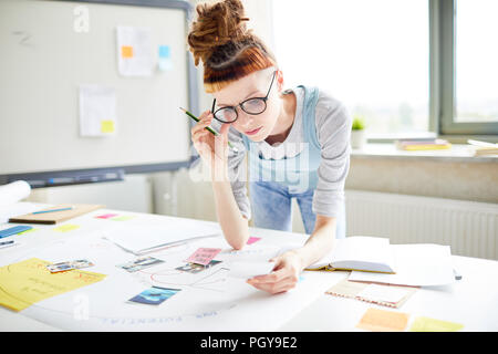 Serious thoughtful female hipster marketer adjusting glasses while reading notes on paper, she learning information necessary for project Stock Photo