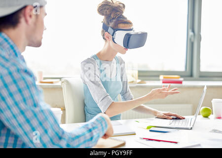 Student startuppers arguing about project: hipster woman with dreads wearing VR headset and pointing at laptop screen while explaining her viewpoint Stock Photo